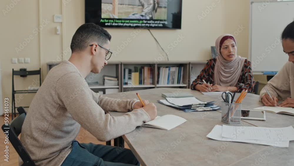 Middle Eastern and Black migrant students learning English language sitting at table in modern classroom writing essays Middle Eastern and Black migrant students learning English language sitting at table in modern classroom writing essays