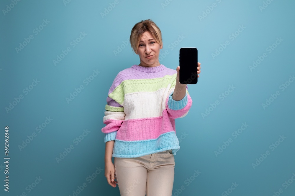 pleasant blond young woman in a striped sweater demonstrates a smartphone mockup on a blue background pleasant blond young woman in a striped sweater demonstrates a smartphone mockup on a blue background