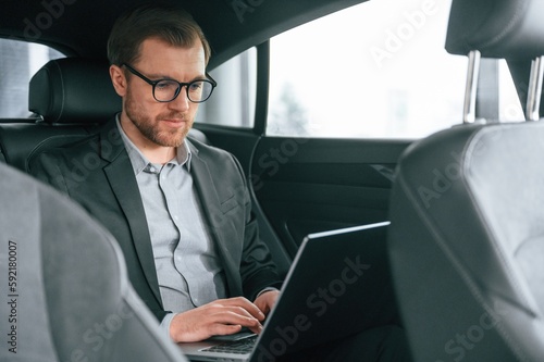 In glasses, working by using laptop. Man in formal business clothes is sitting in the modern automobile