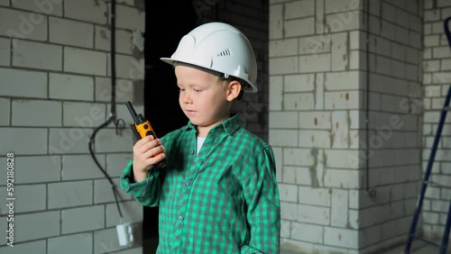 A 5-year-old boy of the buzzer Z generation plays on a construction site in a white helmet and with a walkie-talkie in his hand. A boy in a green shirt is talking on a radio at a site. High quality 4k photo