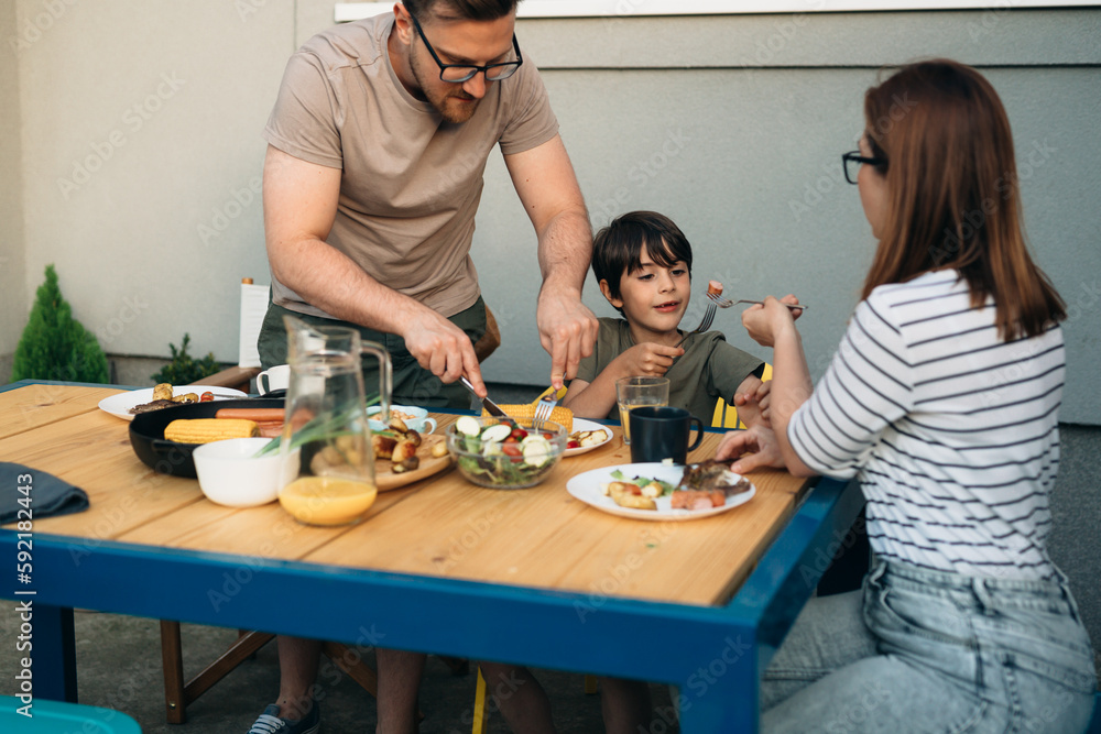 Father cuts meat for his son while mother feeds him Father cuts meat for his son while mother feeds him