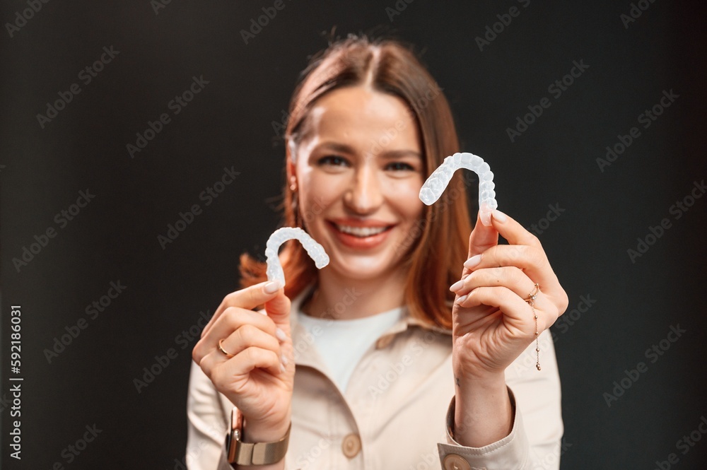 Young woman is standing against black background and holding aligners Young woman is standing against black background and holding aligners