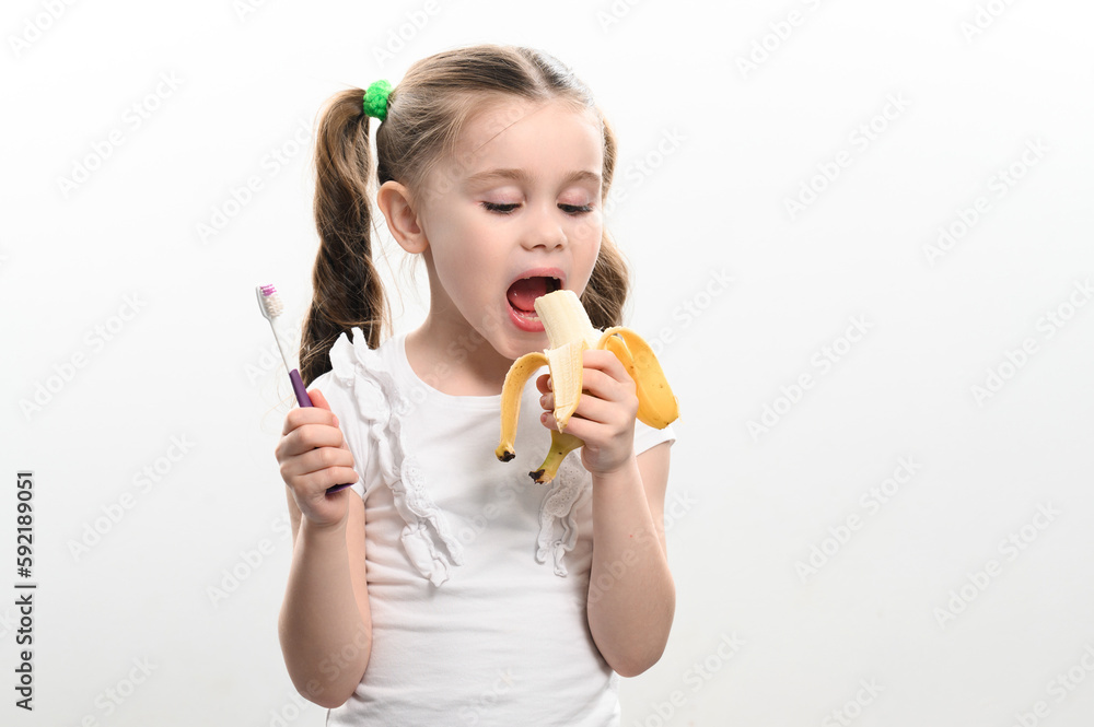 A little girl eats a banana and holds a toothbrush in her hands, studio photo on a white background, a banana helps against caries. A little girl eats a banana and holds a toothbrush in her hands, studio photo on a white background, a banana helps against caries.