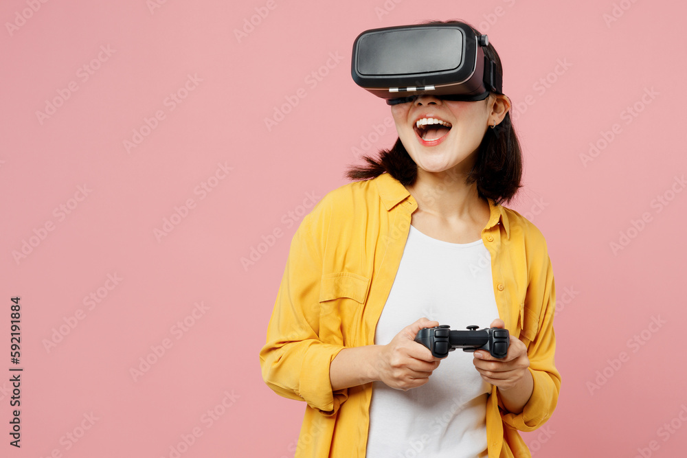 Young excited woman of Asian ethnicity wear yellow shirt white t-shirt hold play pc game with joystick console watching in vr headset pc gadget isolated on plain pastel light pink background studio. Young excited woman of Asian ethnicity wear yellow shirt white t-shirt hold play pc game with joystick console watching in vr headset pc gadget isolated on plain pastel light pink background studio.