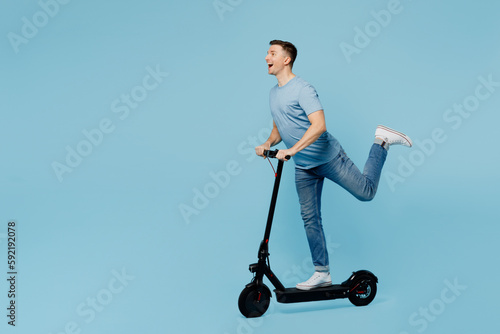 Full body sideways young positive cheerful smiling happy man wears casual t-shirt riding e-scooter look camera isolated on plain pastel light blue cyan background studio portrait. Lifestyle concept.