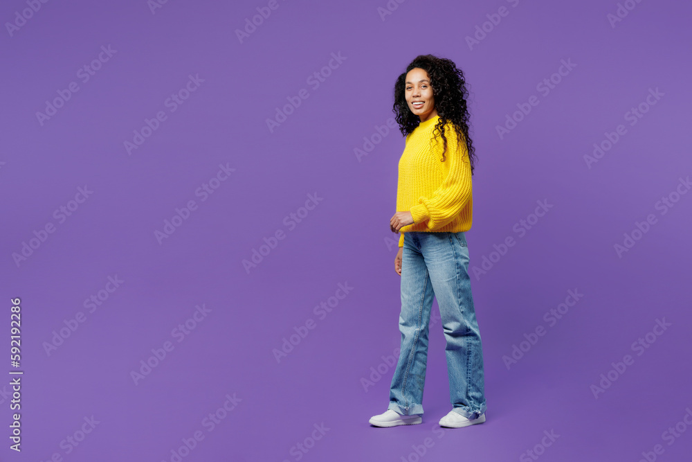 Full body side profile view happy young woman of African American ethnicity wears casual yellow sweater walking going strolling isolated on plain purple background studio portrait Lifestyle concept. Full body side profile view happy young woman of African American ethnicity wears casual yellow sweater walking going strolling isolated on plain purple background studio portrait Lifestyle concept.