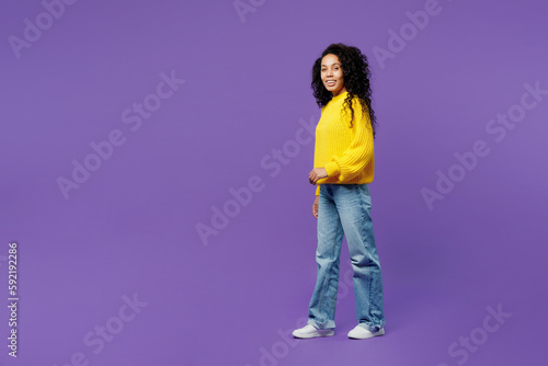 Full body side profile view happy young woman of African American ethnicity wears casual yellow sweater walking going strolling isolated on plain purple background studio portrait Lifestyle concept.