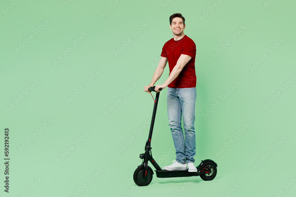 Full body side profile view fun young man he wearing red t-shirt casual clothes riding electric scooter look camera isolated on plain pastel light green background studio portrait. Lifestyle concept. Full body side profile view fun young man he wearing red t-shirt casual clothes riding electric scooter look camera isolated on plain pastel light green background studio portrait. Lifestyle concept.