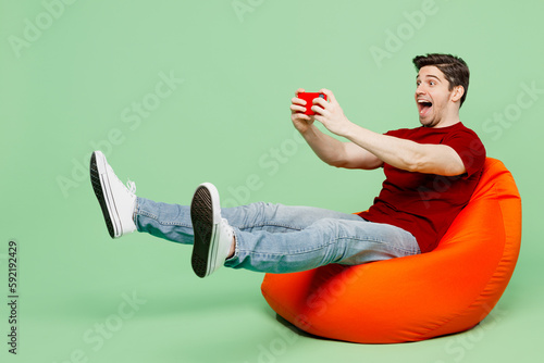 Full body gambling young man wears red t-shirt casual clothes use play racing app on mobile cell phone hold gadget smartphone for pc video games isolated on plain pastel light green background studio.