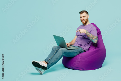 Full body young IT man he wears purple t-shirt sit in bag chair hold use work point finger on laptop pc computer isolated on plain pastel light blue cyan background studio portrait. Lifestyle concept.