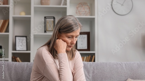Sad senior woman sitting on the sofa sad because of bad news, problems in the family. Experiences due to menopause, health problems, migraines, bad mood.
