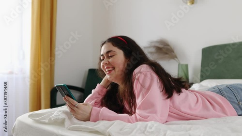 Weekend relaxation. Portrait of young pretty Caucasian woman using smartphone luying on bed. Concept of online leisure and procrastination. photo