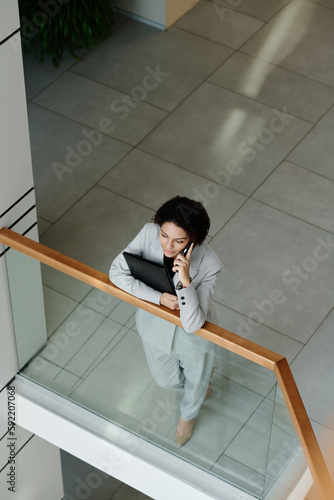 High angle view of young businesswoman in suit talking on mobile phone standing on balcony of office building