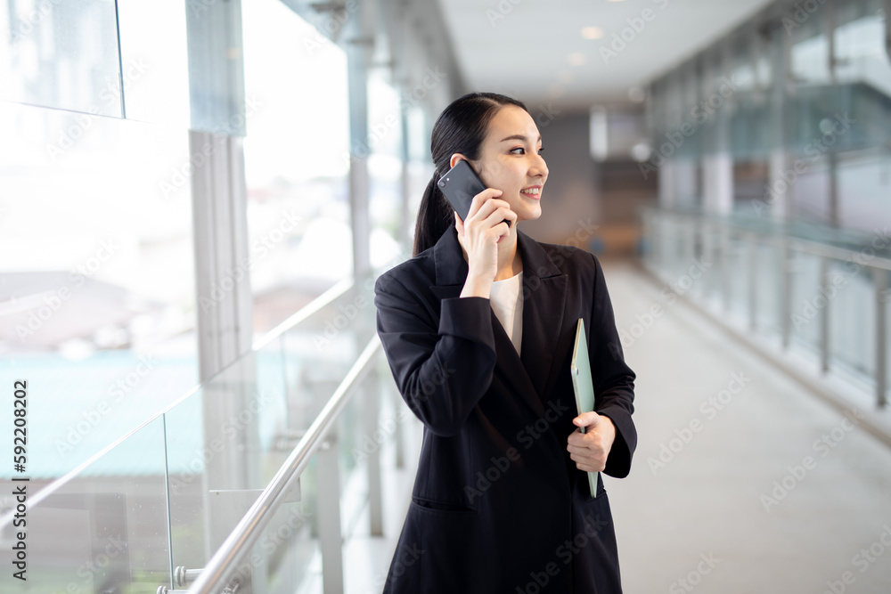 Happy young asian businesswoman talking on the mobile phone and smiling while walking against street blurred building background. Happy young asian businesswoman talking on the mobile phone and smiling while walking against street blurred building background.