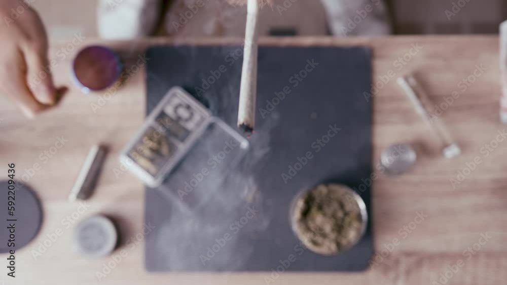 Lighting Up Rolled Marijuana Joint Against Blurred Cannabis Backdrop On The Table. overhead Lighting Up Rolled Marijuana Joint Against Blurred Cannabis Backdrop On The Table. overhead