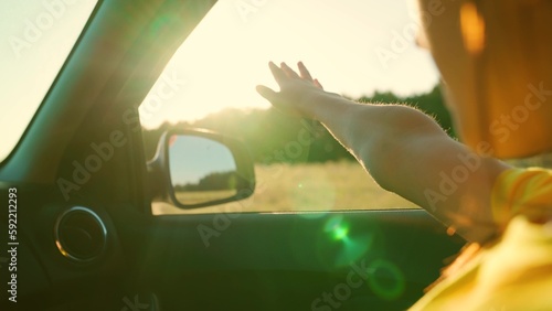 Fototapeta Naklejka Na Ścianę i Meble -  Young woman travels by car catches wind with her hand from car window. Girl with long hair is sitting in front seat of car, stretching her arm out window and catching glare of setting sun. Vacation