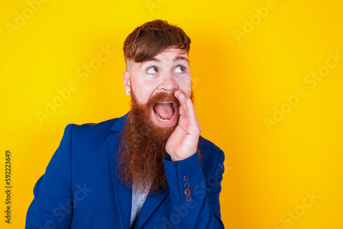 red haired man wearing blue suit over yellow studio background hear incredible private news impressed scream share