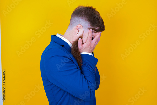 Sad red haired man wearing blue suit over yellow studio background crying covering her face with her hands. © Roquillo