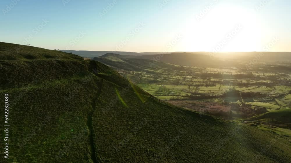 Descending aerial shot with lens flares showing the sunrise lighting up Mam Tor in the peak district, UK with silhouettes of hikers enjoying the view. Descending aerial shot with lens flares showing the sunrise lighting up Mam Tor in the peak district, UK with silhouettes of hikers enjoying the view.