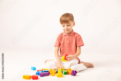 A happy child collects a tower of colored cubes. The preschooler sits down at the white background and assembles the construction set. The boy smiles, pleased with the result.