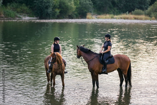 Riders, two young women riding beautiful horses down the calm river surrounded by the green grove
