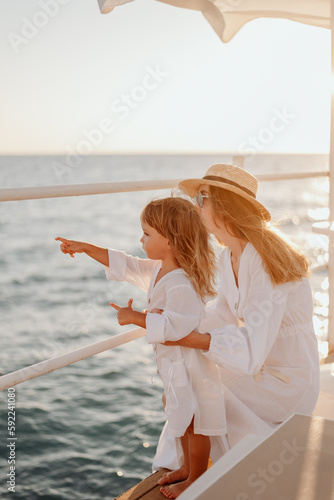 Mom and daughter in white dresses, at sunset, look at the ocean from the pier. Daughter points a finger into the distance