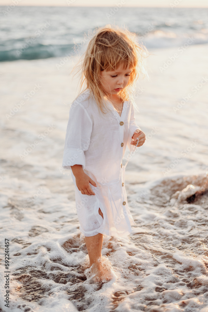 A little girl with long hair, in a white dress, walks barefoot along ...