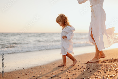 A little girl and a young mother in white dresses and a hat are walking on the water on the ocean at sunset. Deserted beach.