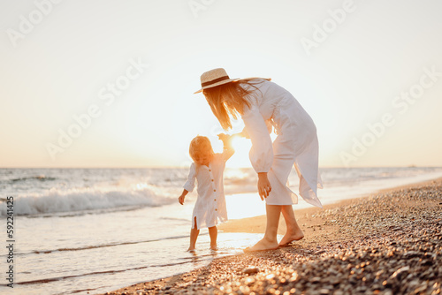 Fotobehang A young mother holds her little daughter by the hand and together they walk along the ocean towards the sunset