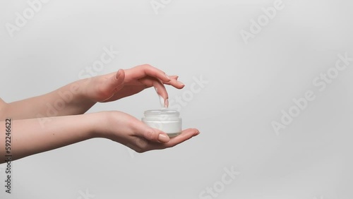Woman is holding glass jar of face cream in her hand, taking a sample with a finger, on gray background, copy space. 4k, slow motion.