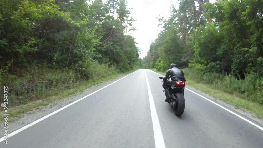 Follow to motorcyclist rides on motorcycle on woodland country road. Man in helmet races modern sport motorbike at rural route near forest. Guy driving bike during trip or journey. Concept of freedom Follow to motorcyclist rides on motorcycle on woodland country road. Man in helmet races modern sport motorbike at rural route near forest. Guy driving bike during trip or journey. Concept of freedom
