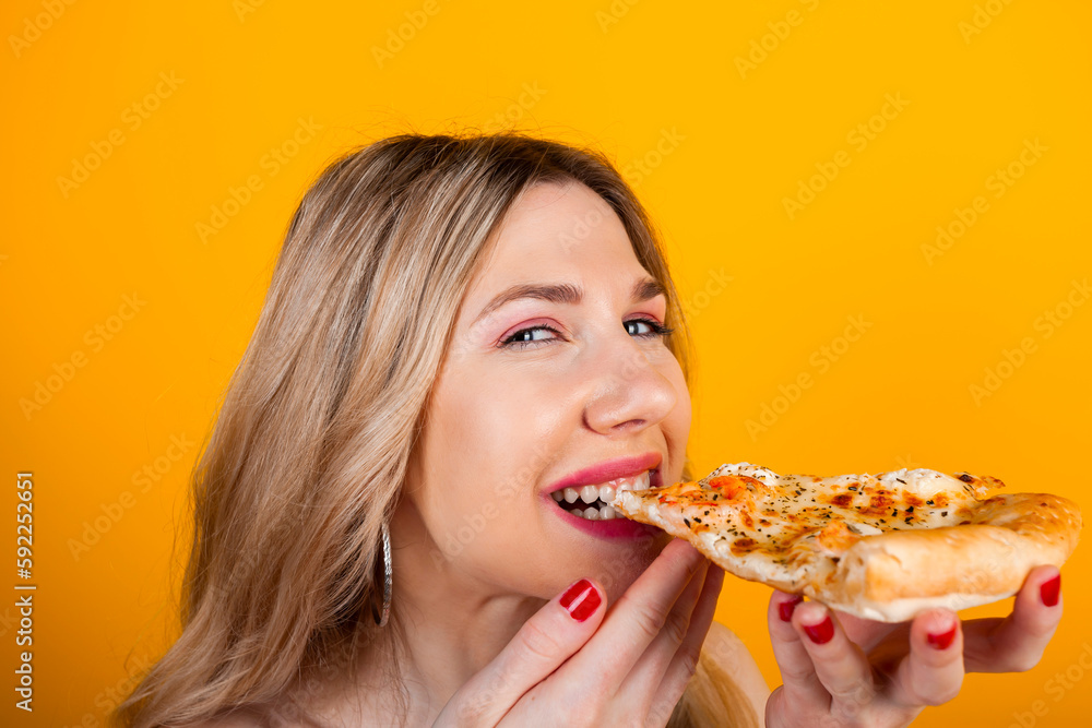 A beautiful fit young blonde girl bites and eating a pizza slice. Isolated on yellow.