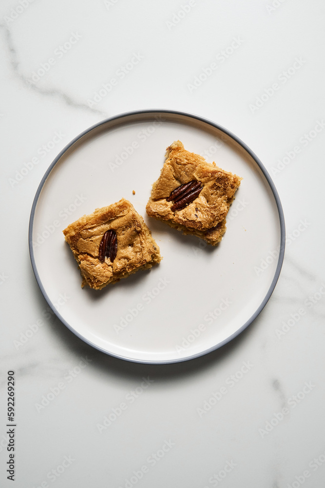 Two squares of Blondie cookies with pecan nuts on white plate. White chocolate sweet and tasty, chewy cookie bars. Top view, copy space Two squares of Blondie cookies with pecan nuts on white plate. White chocolate sweet and tasty, chewy cookie bars. Top view, copy space