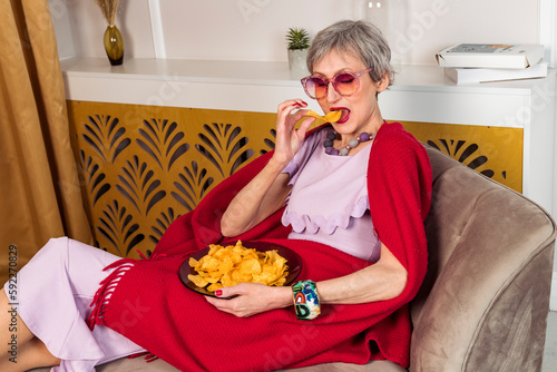 A nice stylish elderly lady in a pink dress and a large scarf is draped over her shoulders is lying on a couch in a modern living room and eating potato chips from a plate.