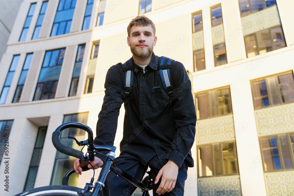 Portrait of a bicycle courier outside. A pizza deliveryman on a bicycle looks at the camera against the background of city buildings Portrait of a bicycle courier outside. A pizza deliveryman on a bicycle looks at the camera against the background of city buildings