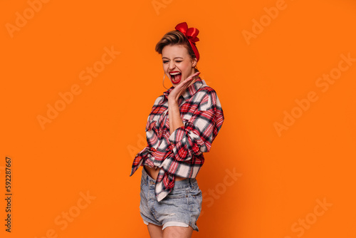 Fashion photo of happy, beautiful woman in short jeans and checkered shirt. Girl laughing, posing on orange studio background.