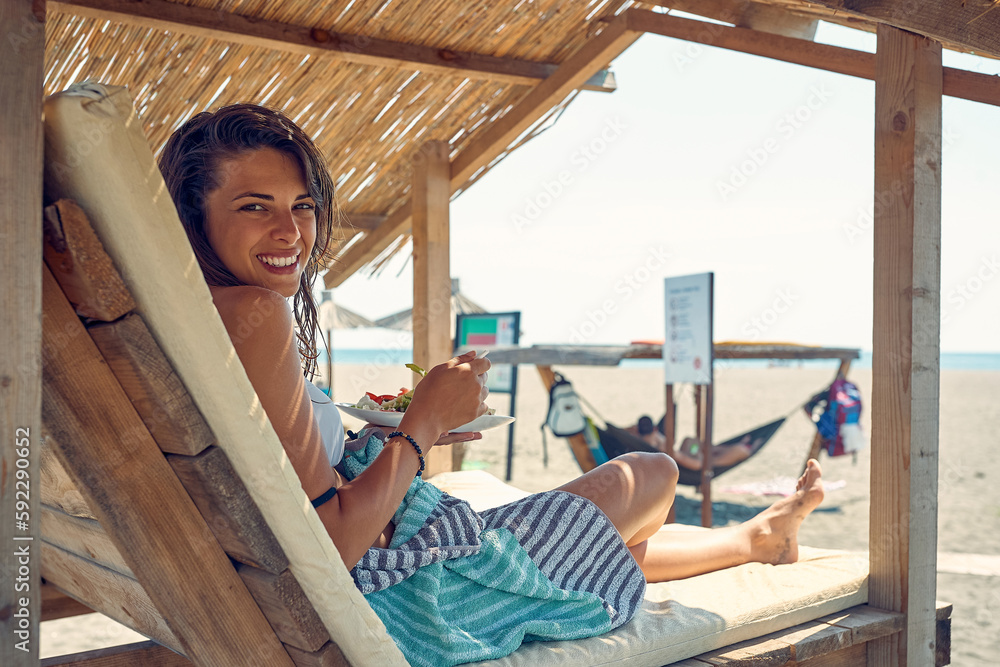 Beautiful brunette woman enjoying healthy meal on beach after swimming. Woman enjoying summer holiday. Travel, lesiure, lifestyle concept.