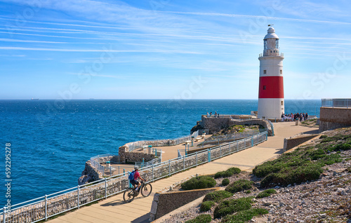 happy senior woman cycling with her electric mountain bike below the Lighthouse of Gibraltar, marking the Strait of Gibraktar, Andalusia, Spain and UK