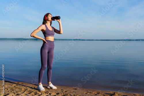 A young woman in a tracksuit drinks water after a running workout on the beach