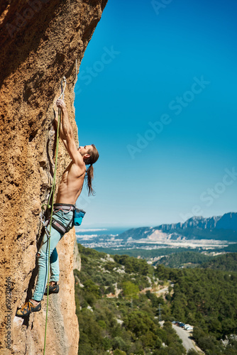 Vertical image athletic male climbing on the high rock with amazing mountains and blue sky behind