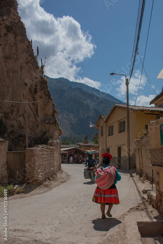 Adult woman with her back turned walking down the street in typical Peruvian dress, and tuc-tuc crossing the street in the city of Ollantaytambo, Peru. 