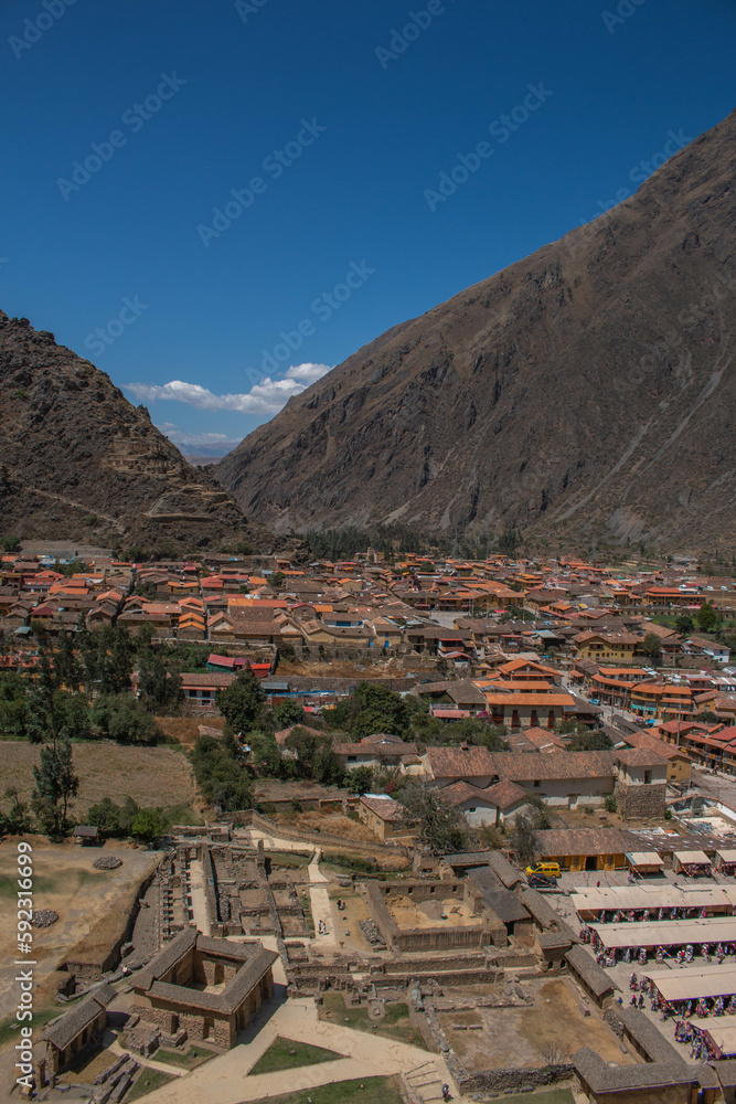 View of the ruins and the city of Ollantaytambo, with the mountains in the background and a blue sky. 