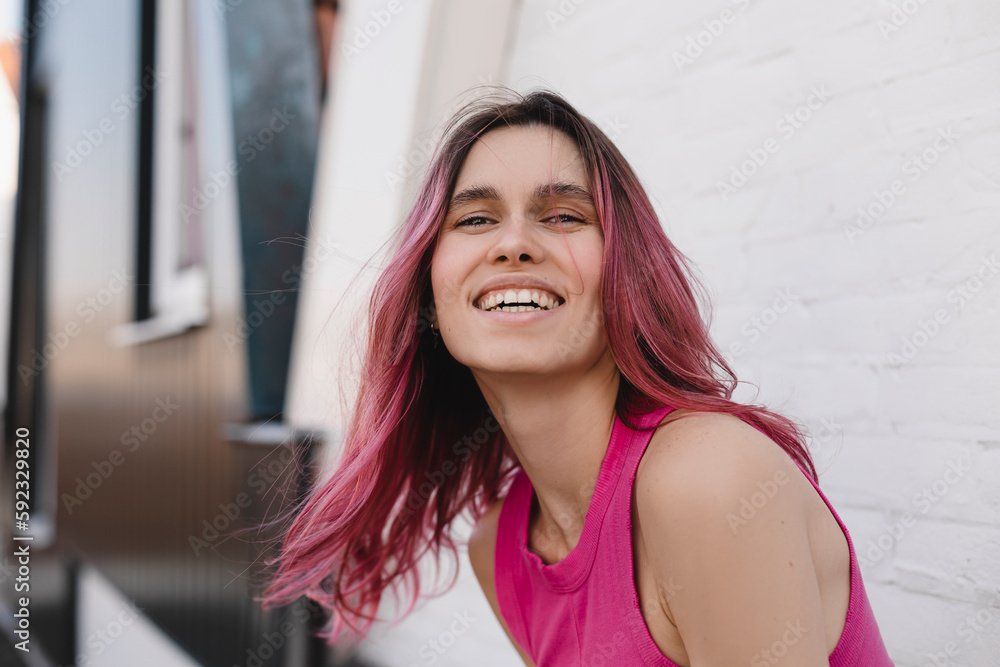 Positive pink hair woman in pink top. Portrait of happy attractive lady at background of white brick wall. Teeth smile woman look at camera, flying hair on the wind.