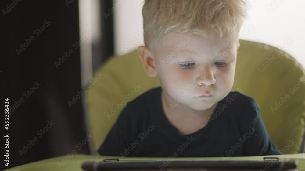 Calm toddler boy watches cartoons via tablet computer sitting in high chair closeup. Blond little child with digital gadget at home. Internet technology Calm toddler boy watches cartoons via tablet computer sitting in high chair closeup. Blond little child with digital gadget at home. Internet technology