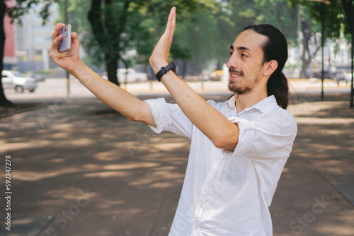 Young Latin man with long hair waving through his cell phone while making a video call from the street.