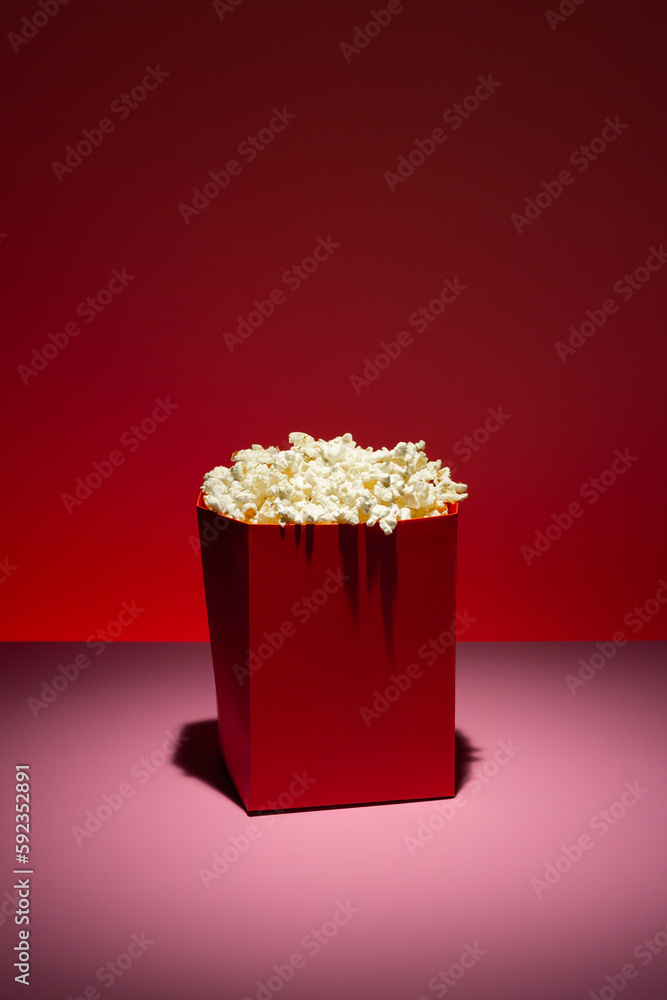Red paper bowl filled with popcorn on pink surface and red gradient background.