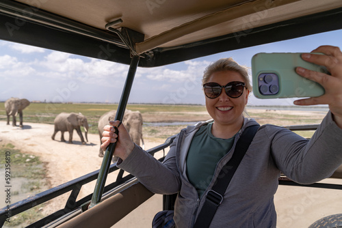 Blonde woman tourist on safari takes a selfie from the vehicle as a herd of elephants passes by