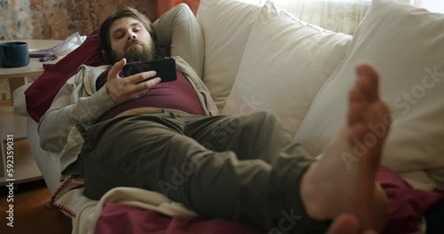 Carefree, careless lazy beard man lying on the couch napping, falling asleep, watching smartphone. Unkept, shabby look. Waste time and motivation problem. Laziness and procrastination concept