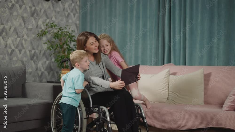 Cute children snuggle up to mom in wheelchair to make selfie on modern tablet. Woman with disability has fun with son and daughter in living room Cute children snuggle up to mom in wheelchair to make selfie on modern tablet. Woman with disability has fun with son and daughter in living room