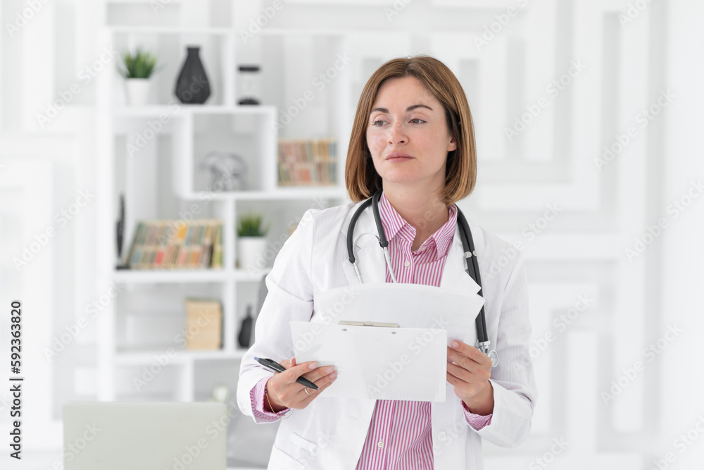 Portrait photo of young and beautiful smiling female doctor in white medical gown standing in the office of the modern clinic